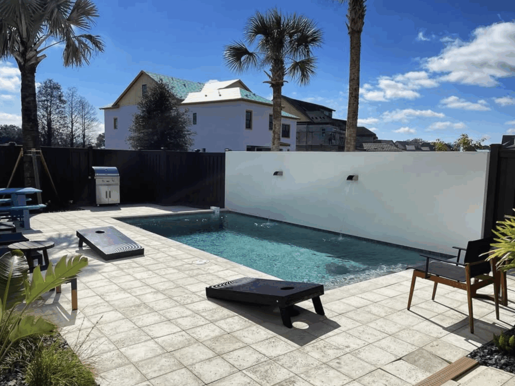 A small outdoor swimming pool with a tiled patio, cornhole boards, a barbecue grill, palm trees, and a white privacy wall with water features.